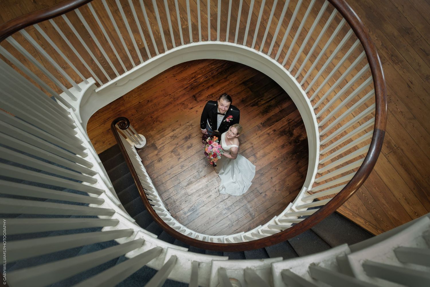 Aerial view of bride and groom embracing on hardwood floor beneath elegant spiral staircase.