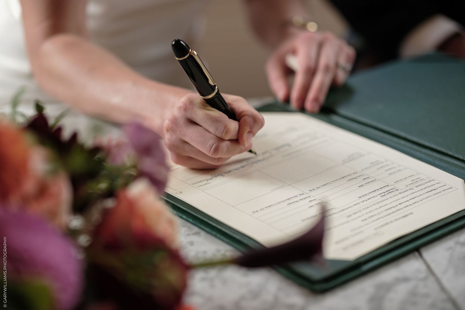 Bride signing wedding certificate with fountain pen beside colorful bridal bouquet at marriage ceremony.