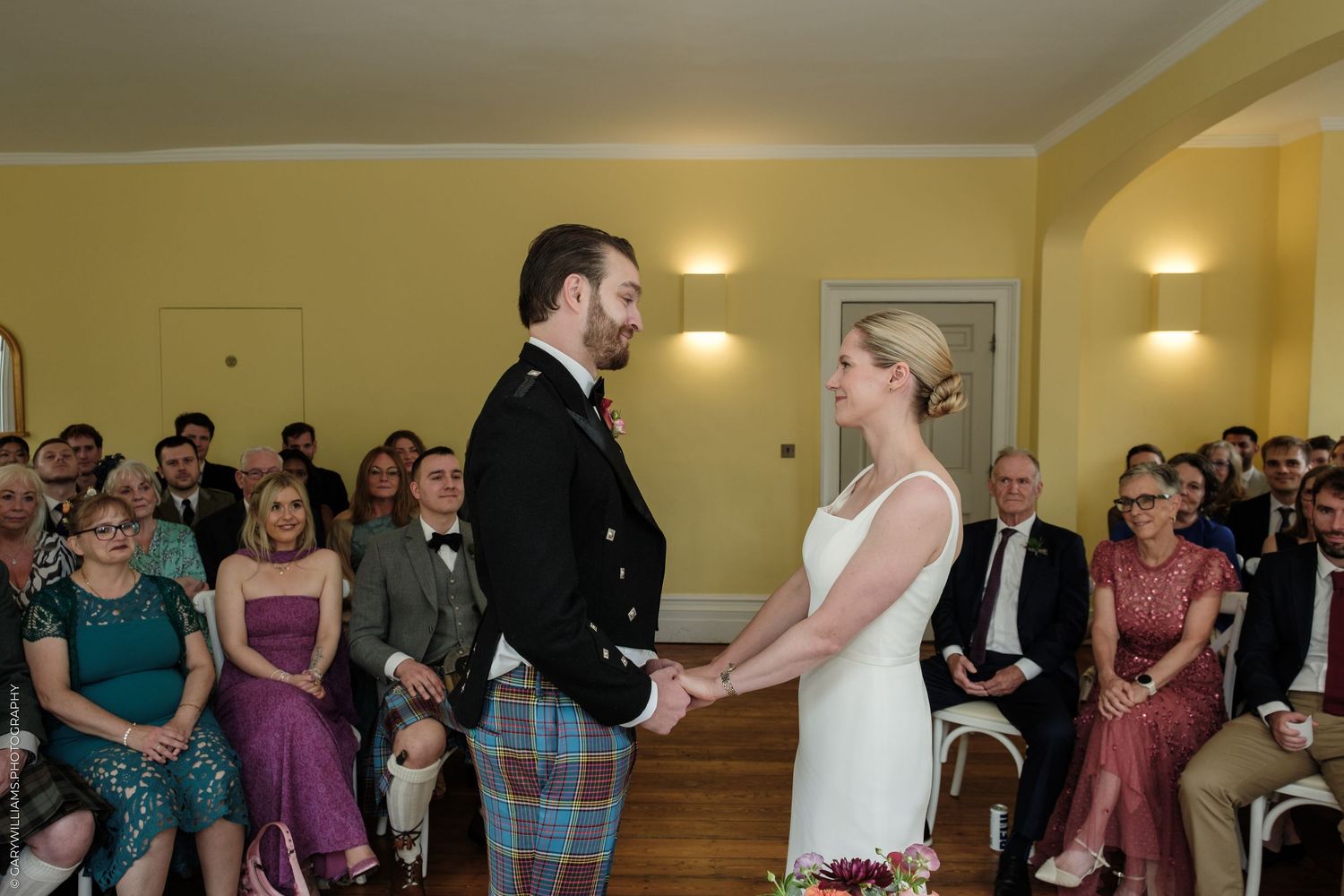 Couple exchanging vows at a wedding ceremony, groom in Scottish tartan kilt, bride in white dress, guests seated behind.