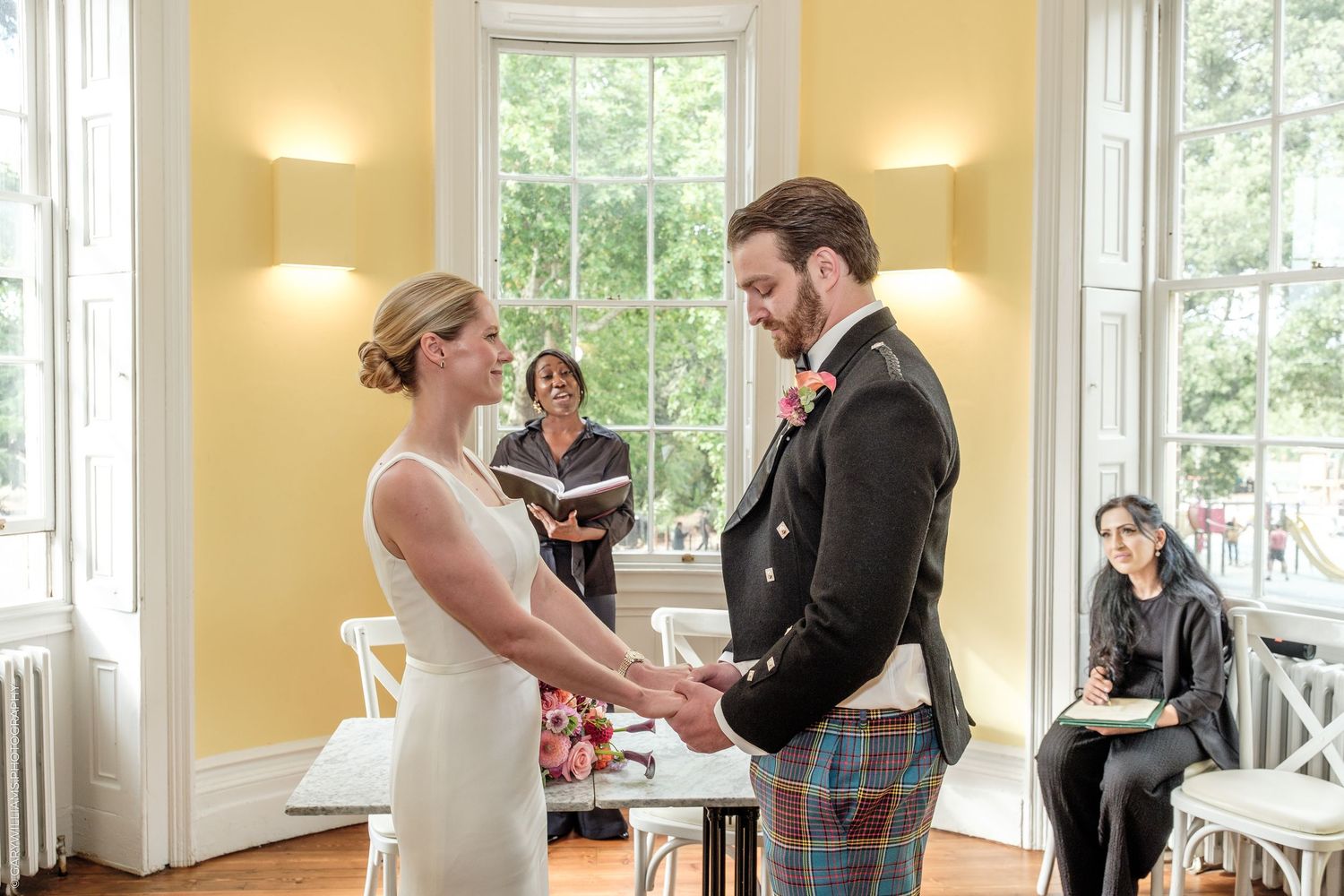 Bride and groom holding hands during indoor wedding ceremony, officiant behind them, yellow walls, bright windows.