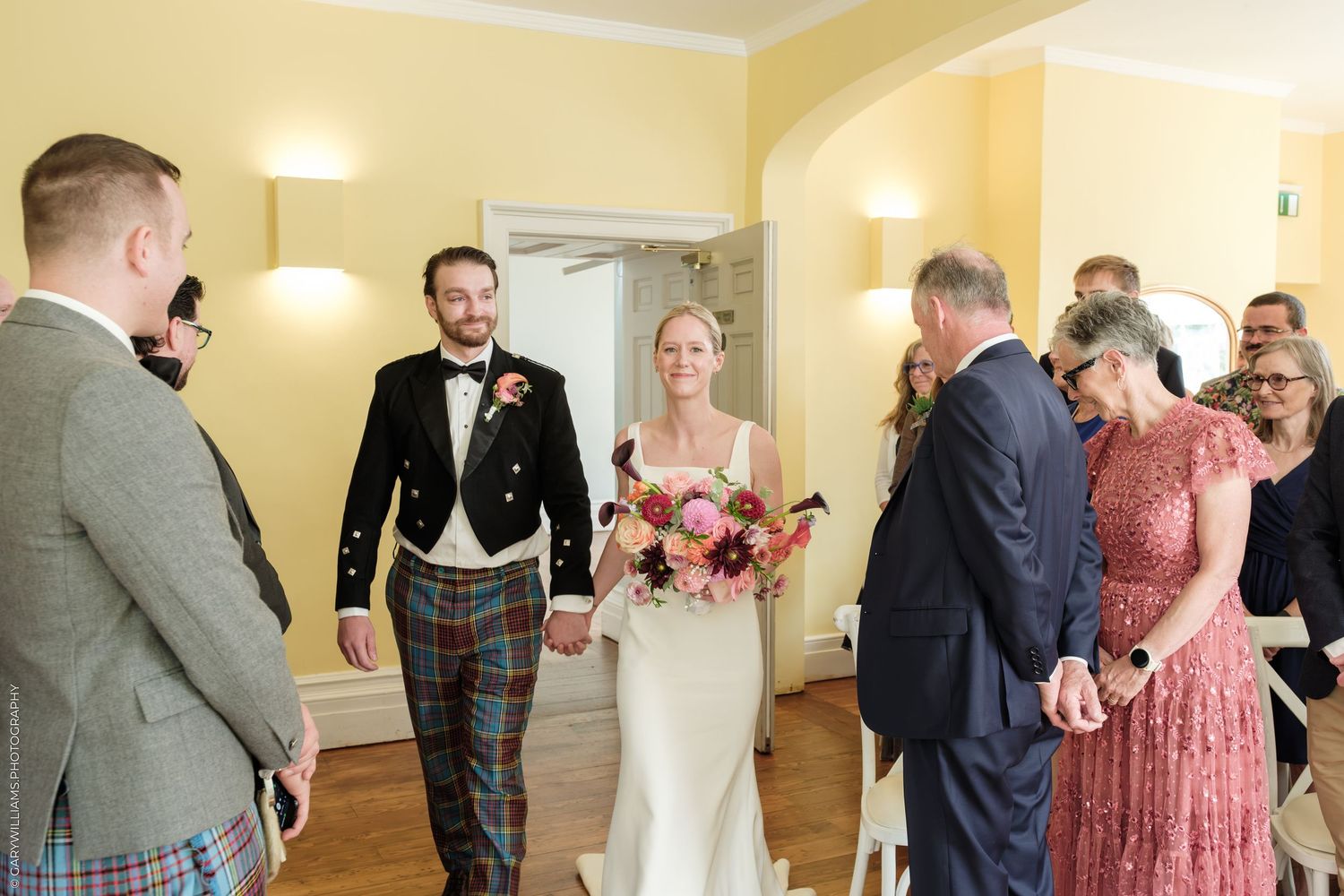 Bride and groom walk down the aisle in a yellow-walled venue, groom wearing tartan kilt, bride holding colorful bouquet.