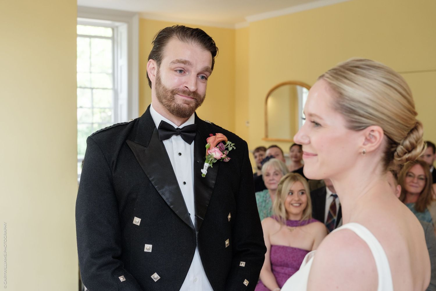 Groom in black tuxedo smiling at bride during wedding ceremony in elegant yellow room.