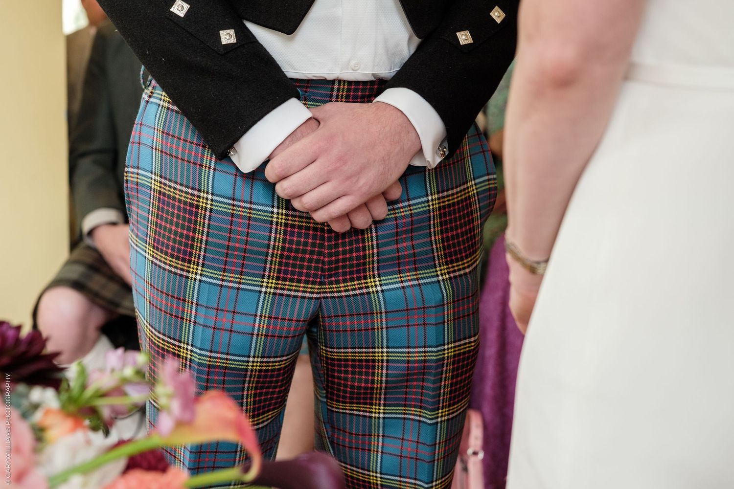 Groom wearing colorful blue tartan plaid trousers at a wedding ceremony, standing beside bride in white dress.
