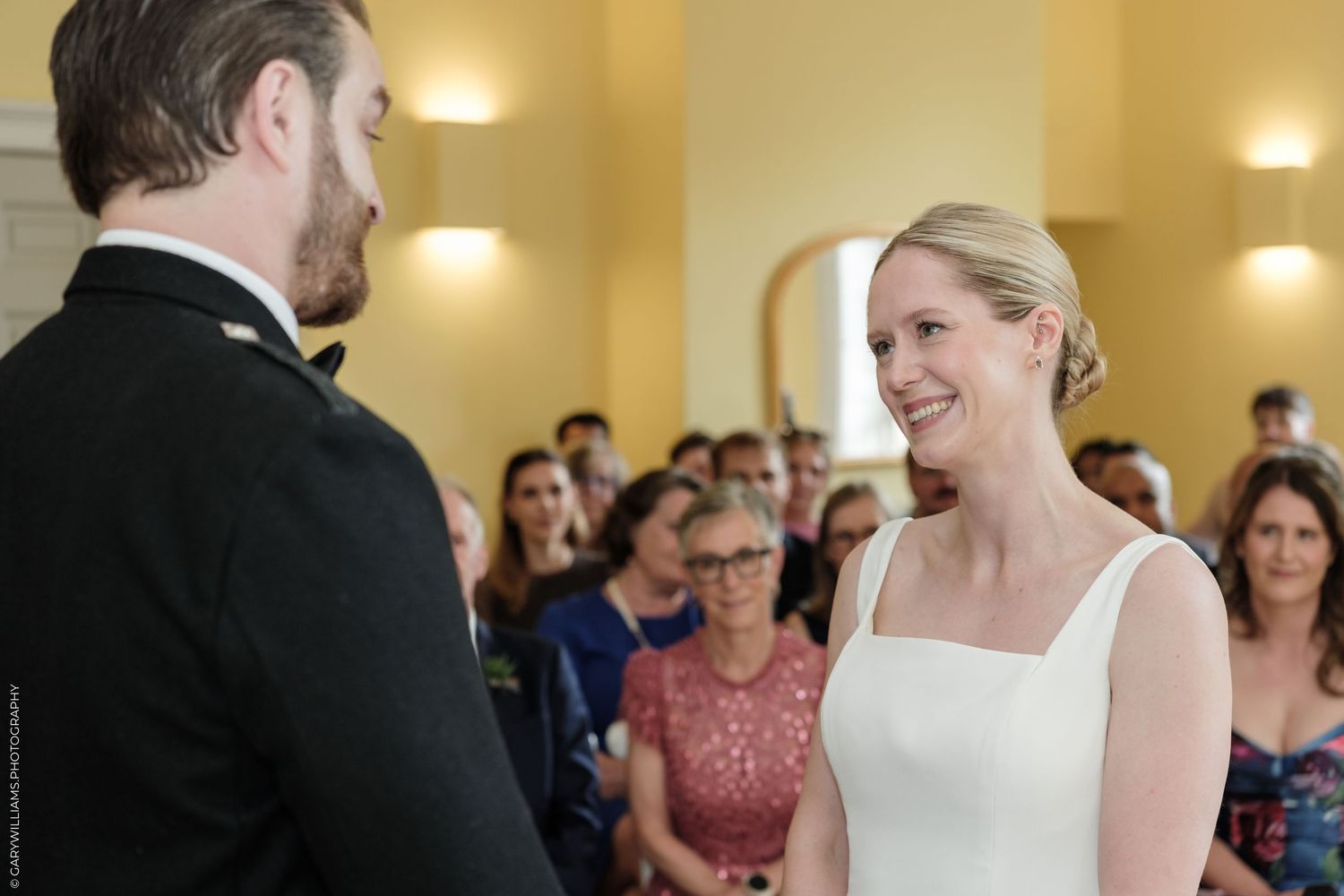Bride smiling at groom during wedding ceremony with guests seated in background.