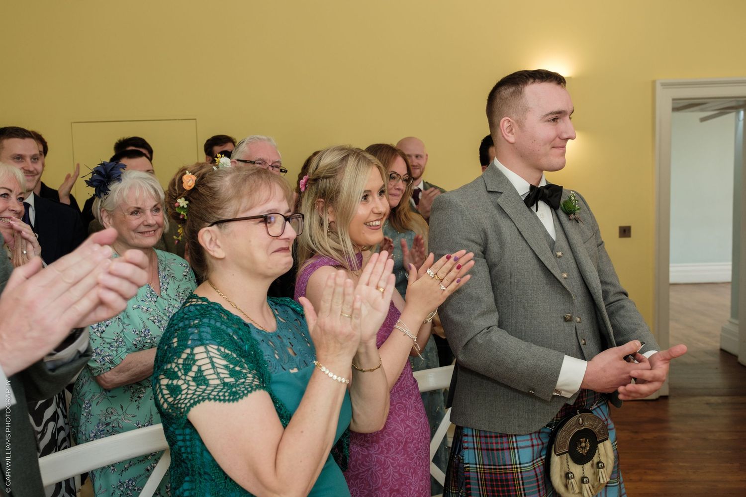 Wedding guests in formal attire clapping and celebrating, man in traditional Scottish kilt standing proudly at ceremony.