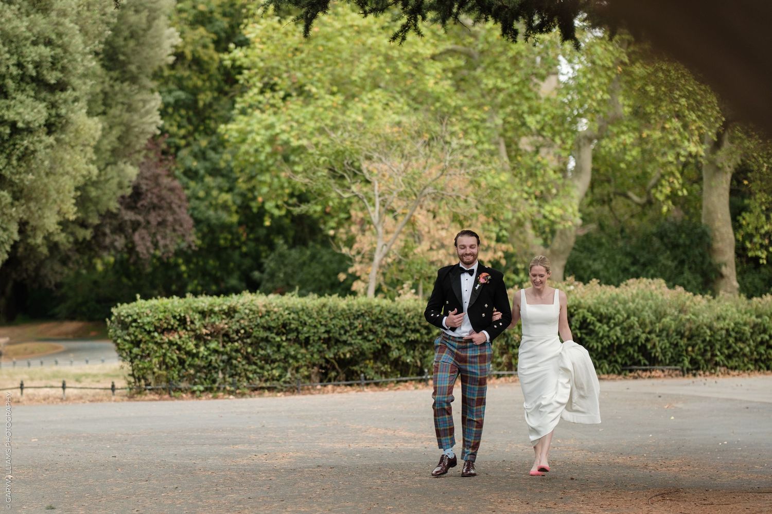 Bride and groom walking hand in hand outdoors in a lush green park, groom wearing traditional Scottish tartan kilt.