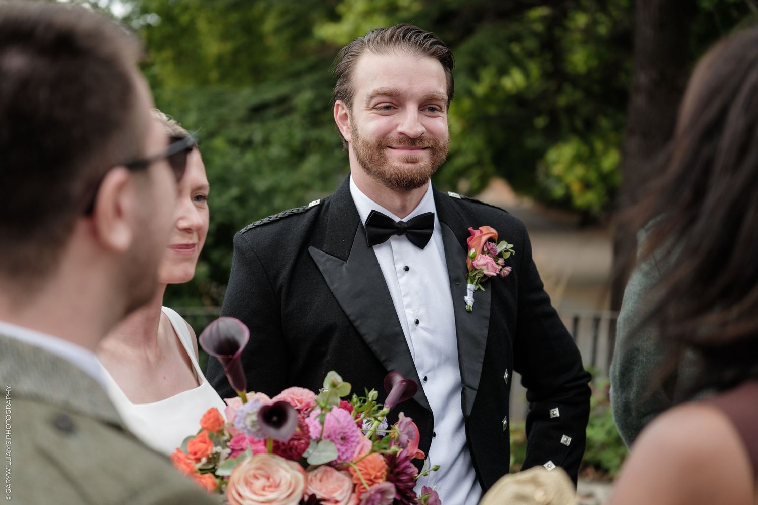 Smiling groom in black tuxedo and bow tie holding colorful bridal bouquet during outdoor wedding ceremony.