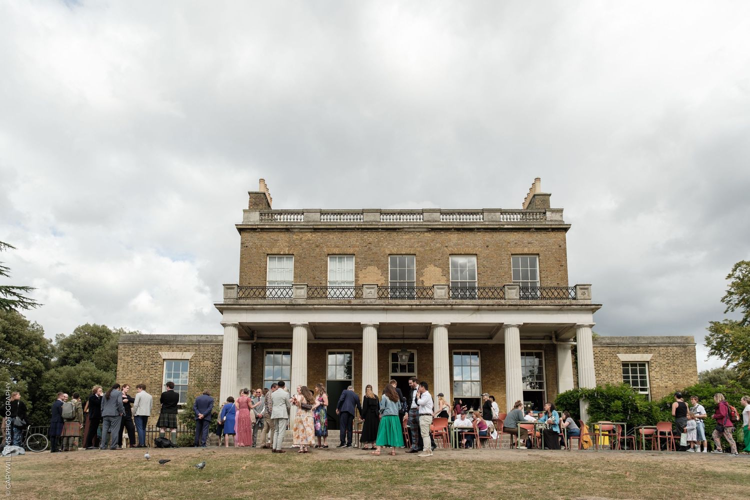 Guests gathering outside elegant Georgian manor house under cloudy sky.