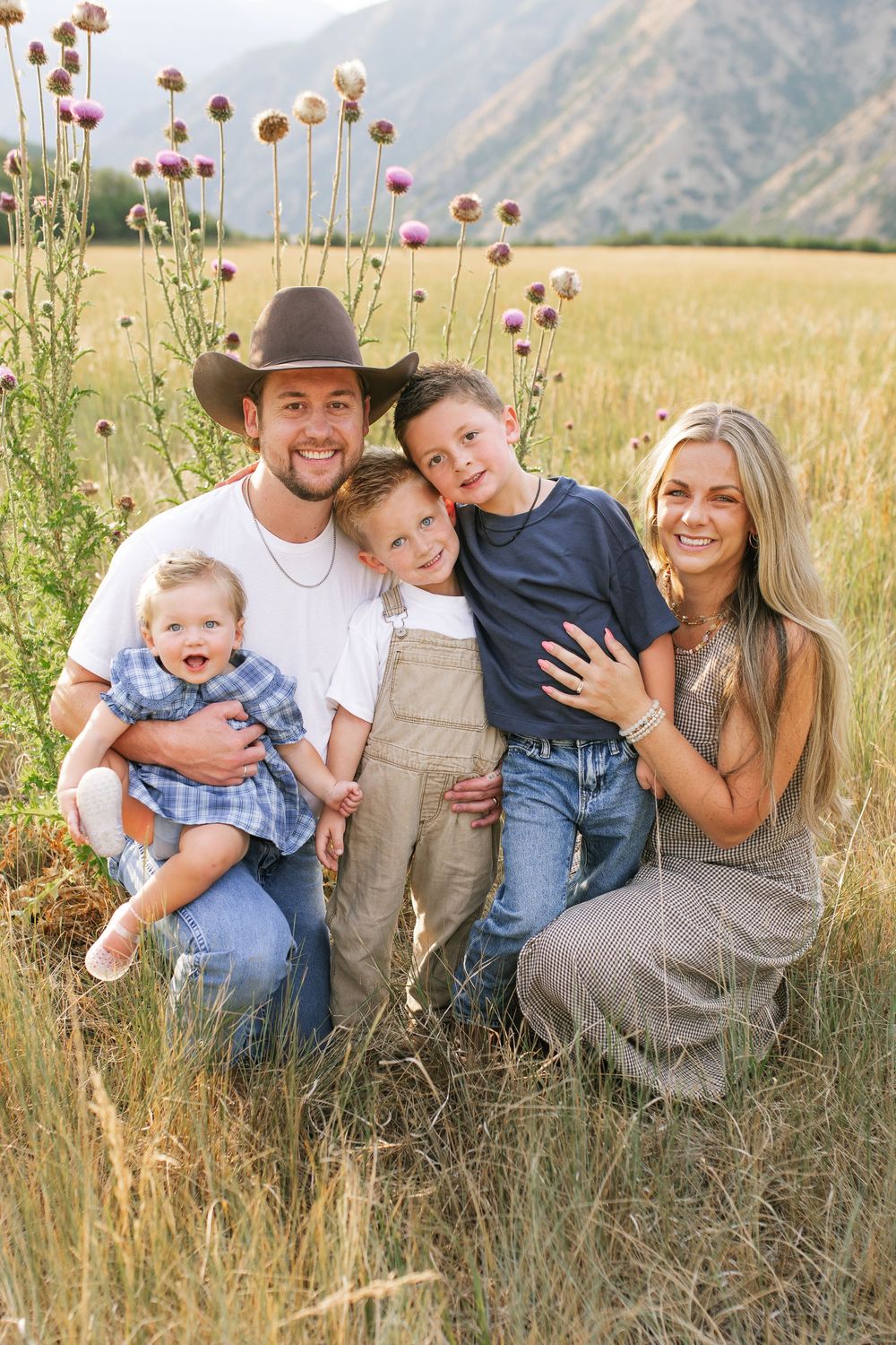 Happy family of five posing in a golden field with mountains, father in cowboy hat holding baby, two young boys and smiling mother.