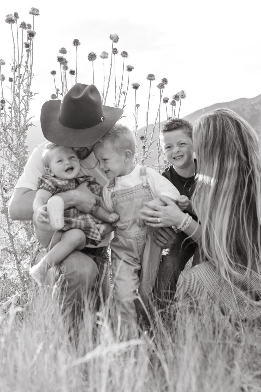 Black and white family photo of parents with three young children laughing in a field, father wearing a cowboy hat.