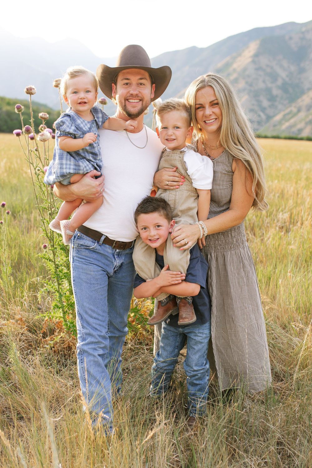 Happy family of five posing in a golden field with mountain backdrop, parents holding three young boys.