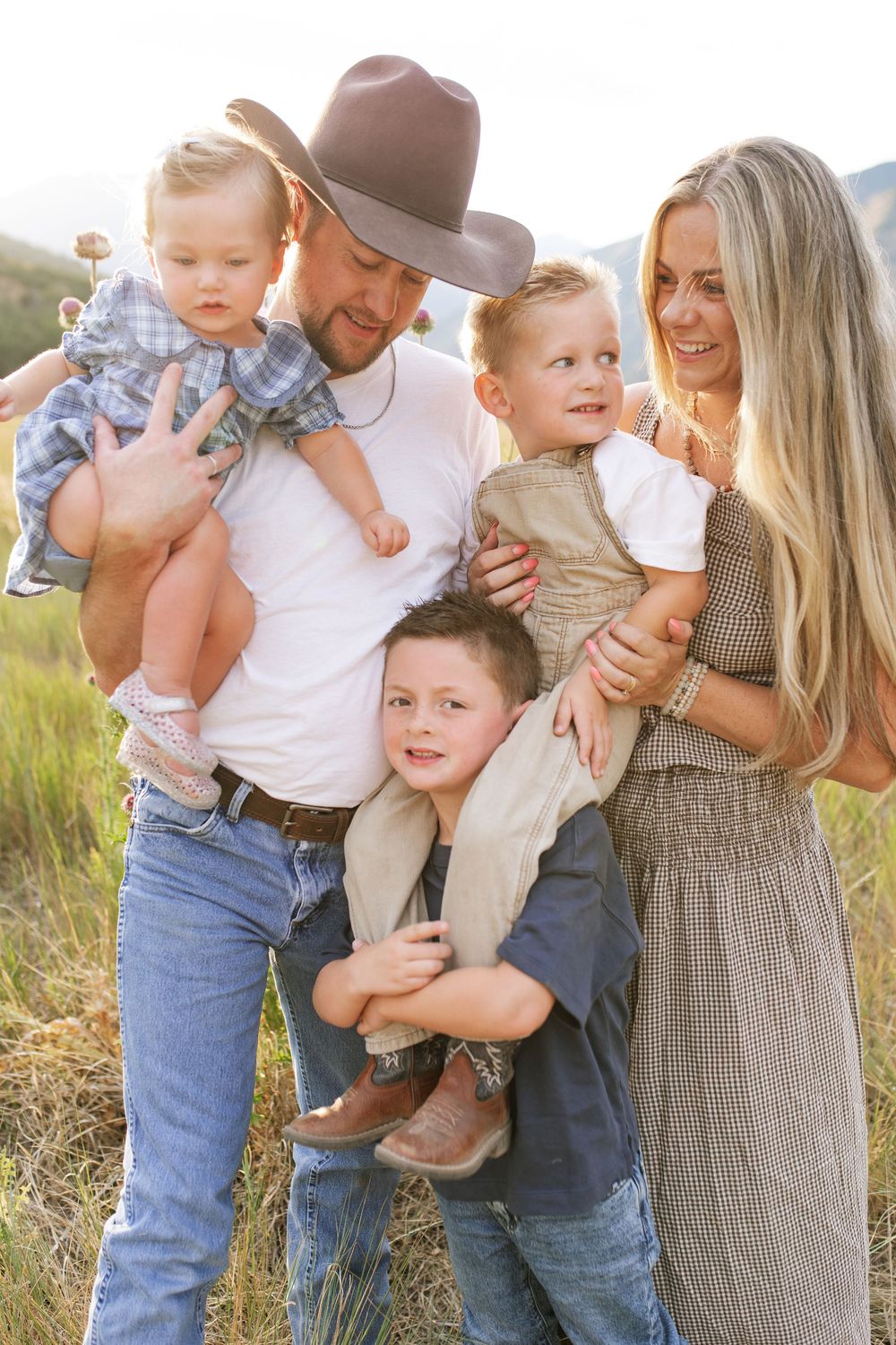 Happy family of five posing outdoors in a sunny field, father in cowboy hat holding toddler, mother with two young boys.
