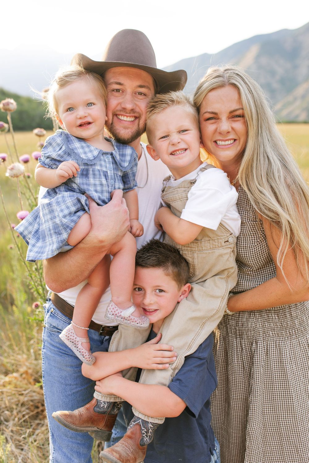 Happy family of five posing outdoors in a mountain meadow, parents holding young children, man wearing cowboy hat.