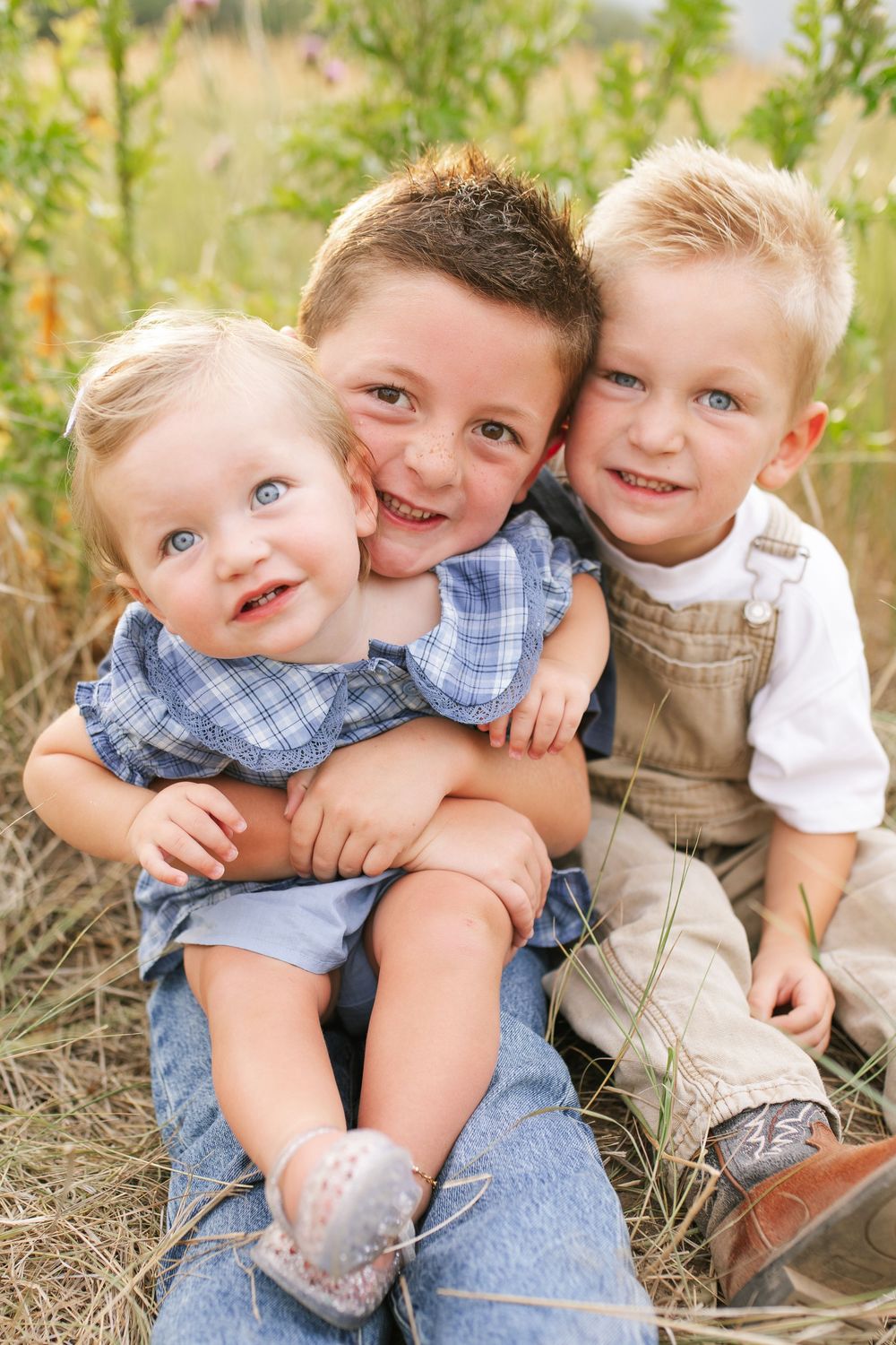 Three young siblings sit together in a field, older dark-haired boy hugging toddler sister and blonde brother.