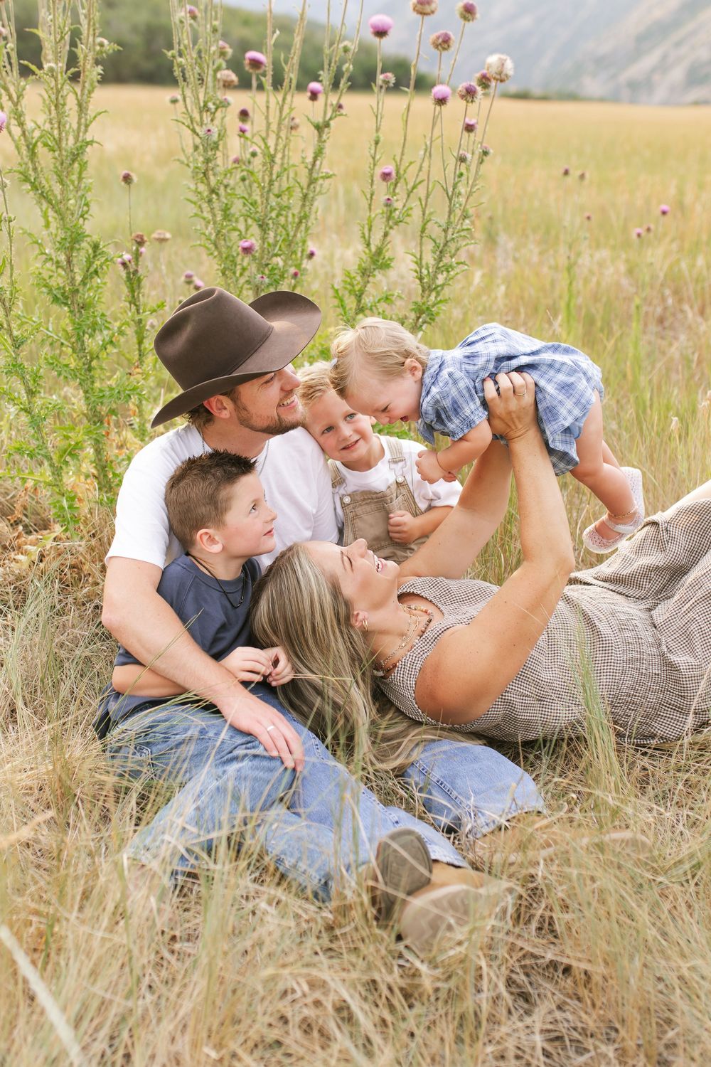 Family of four relaxing in a golden meadow with wildflowers and mountains, parents kissing children outdoors.
