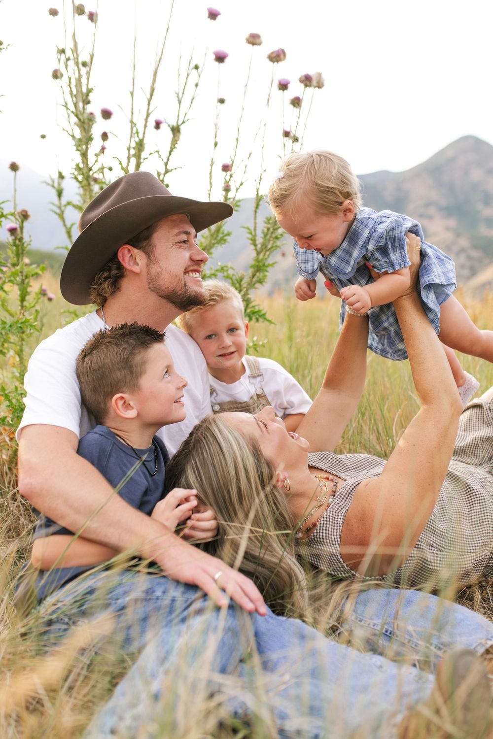 Happy family relaxing in a golden field with mountains, dad in cowboy hat holding toddler while mom laughs with two boys.