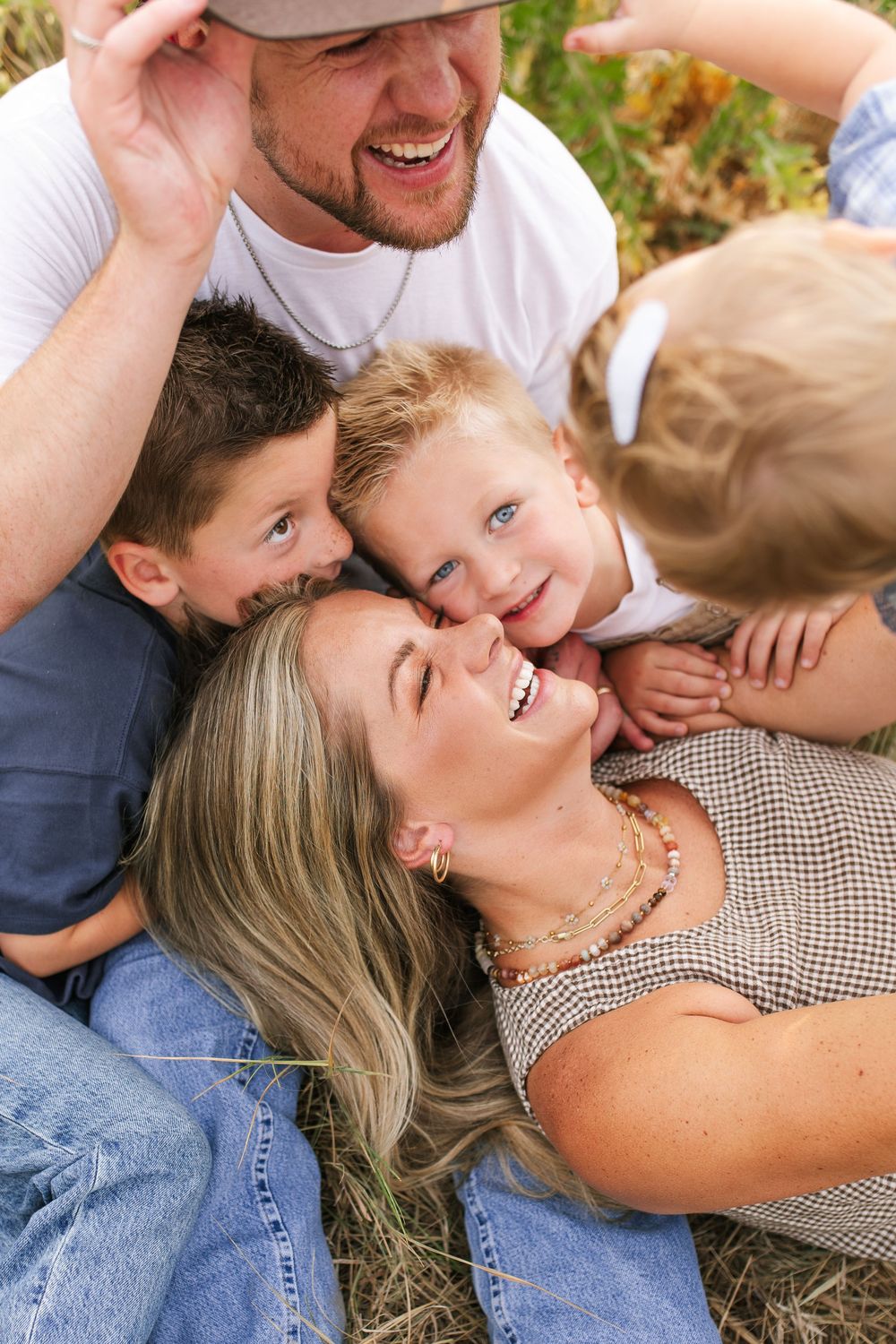 Joyful family portrait with laughing mom lying in grass surrounded by dad and three young children in a warm outdoor setting.