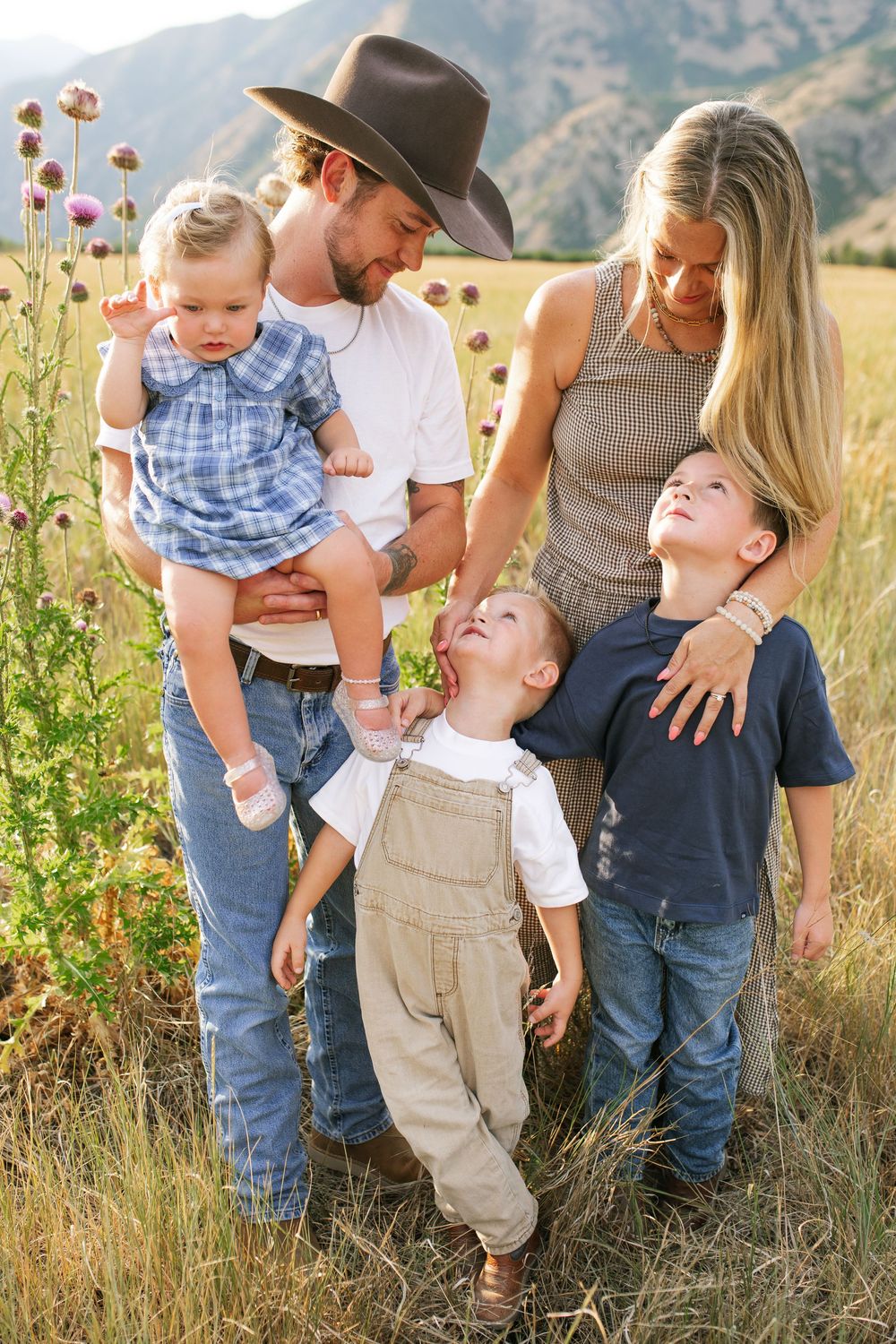 A happy family of five poses in a sunny field with mountain backdrop, dad in cowboy hat holding baby girl.