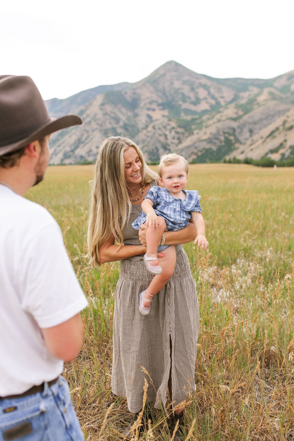 Smiling mother holding toddler in a mountain meadow while father in cowboy hat looks on lovingly.
