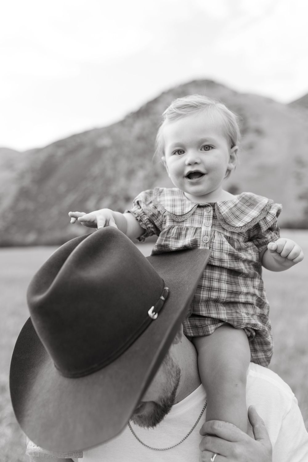 Black and white photo of a happy baby in plaid outfit held up by a man with cowboy hat, mountains in background.
