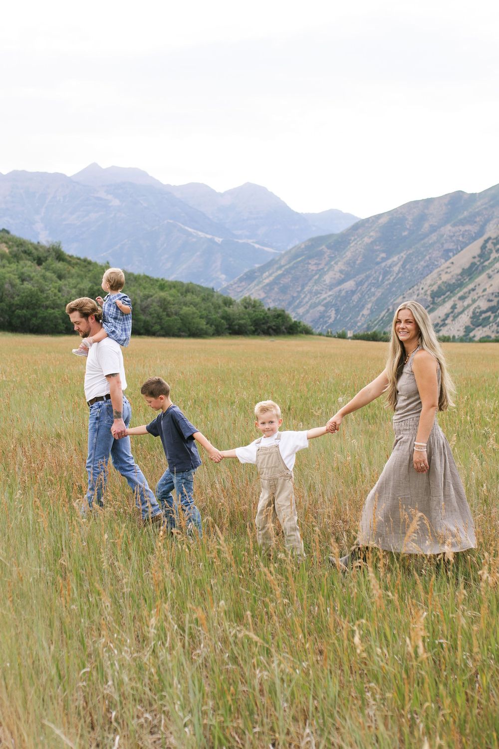 Family of five walking hand-in-hand through tall grass field with scenic mountain backdrop.