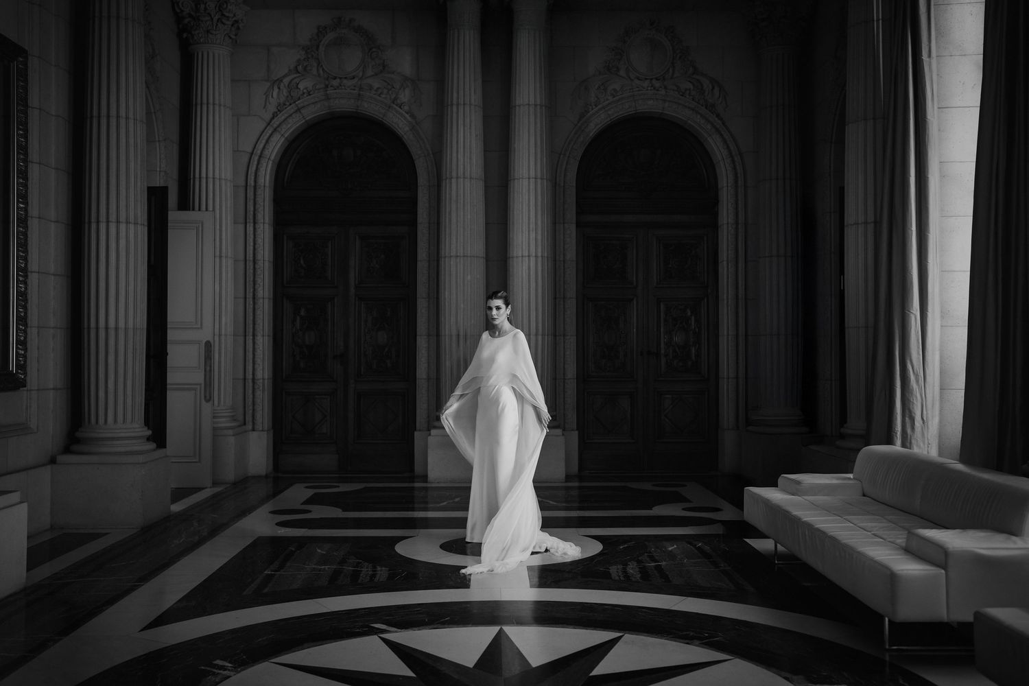Elegant bride in white gown and veil stands in grand black and white marble hall with arched doors.