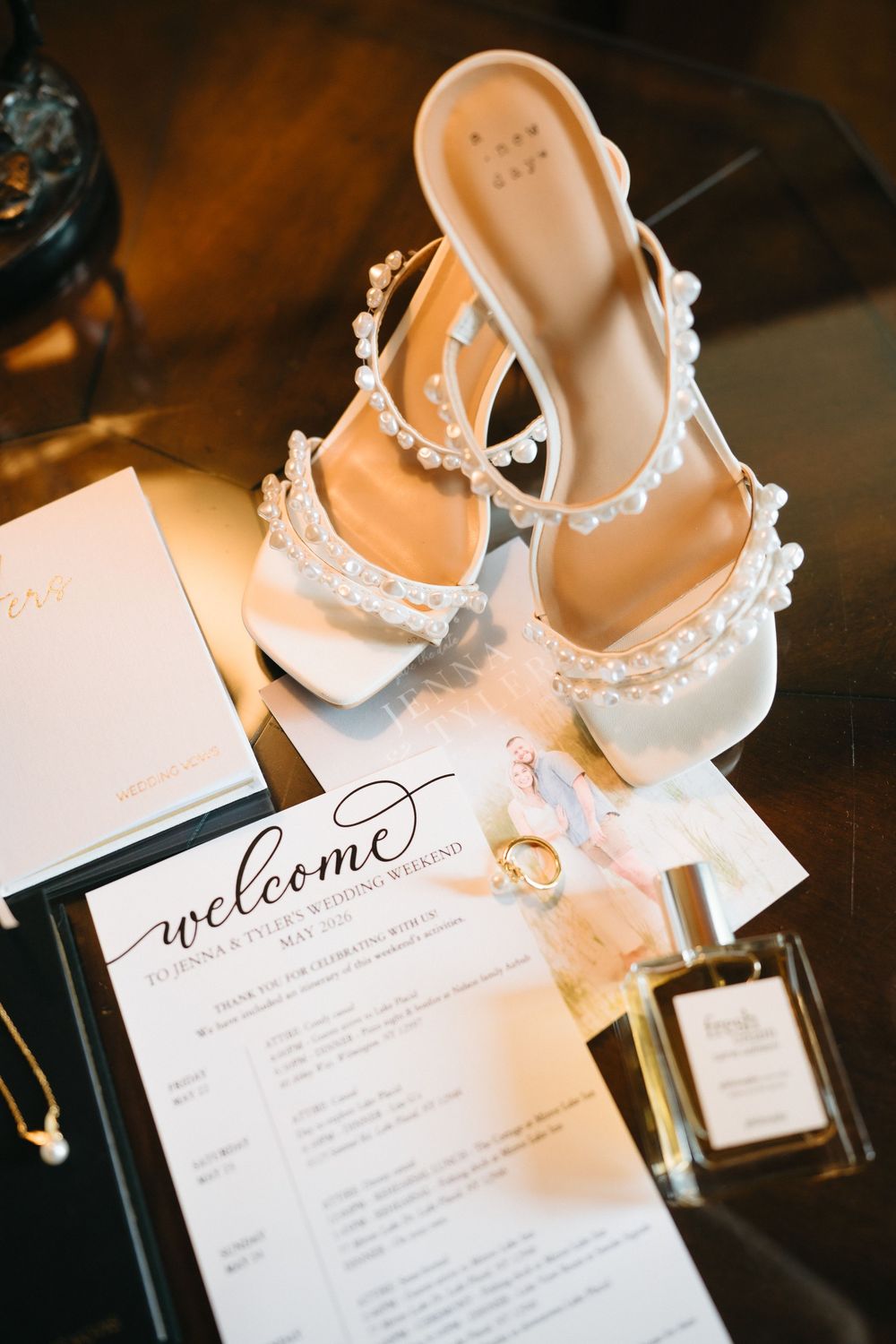 White pearl-embellished bridal heels displayed with wedding rings, perfume, and a welcome itinerary card on a dark surface.