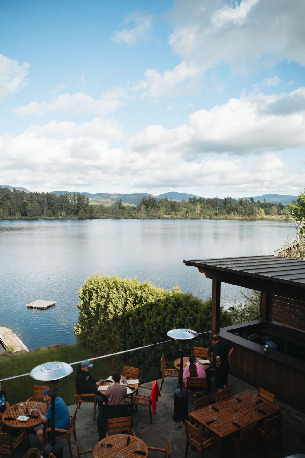 Outdoor lakeside patio with wooden tables and blue umbrellas overlooking a serene lake surrounded by trees and mountains.