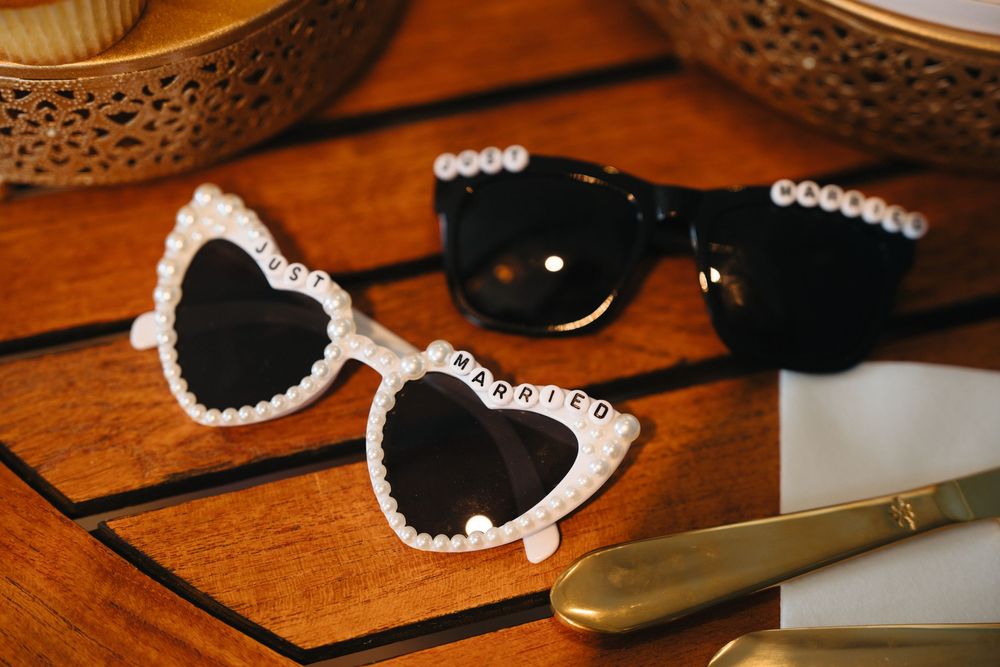 Just Married heart-shaped sunglasses with pearl embellishments beside black sunglasses on a wooden table.