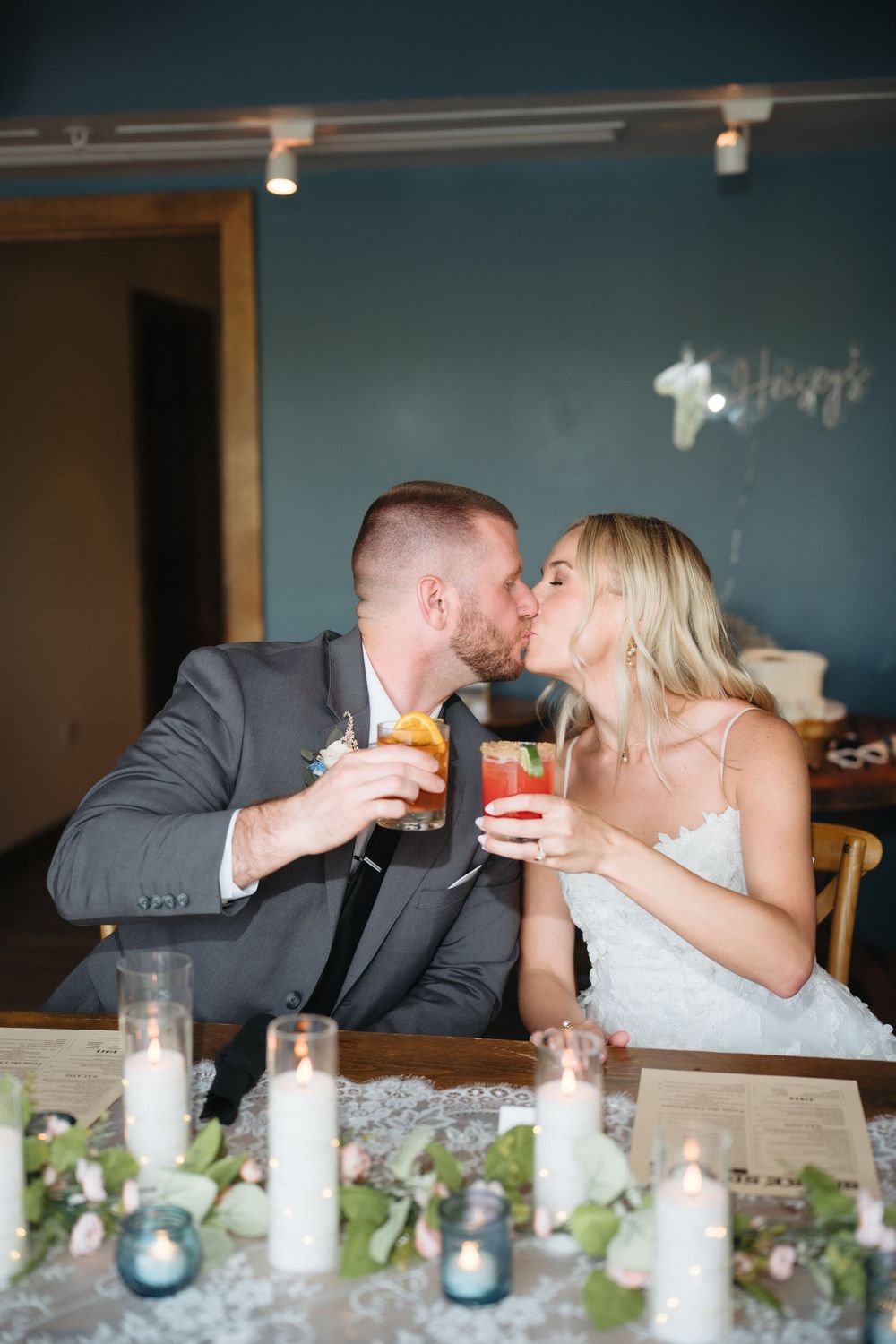 Bride and groom kissing while holding cocktails at decorated wedding reception table with candles and greenery.