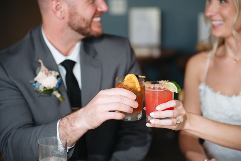 Bride and groom clinking colorful cocktails together, dressed in wedding attire with a floral boutonniere.