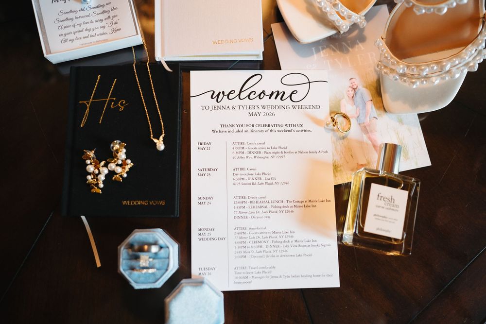 Wedding flat lay with welcome itinerary, gold jewelry, perfume bottle, ring box, and His vow book on dark wood.