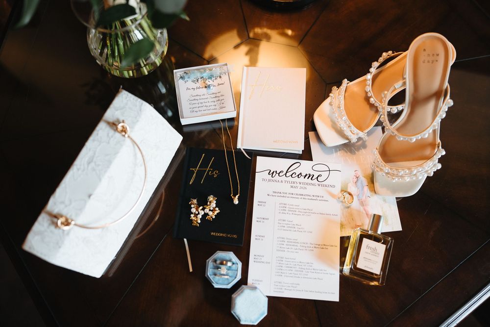 Elegant wedding flat lay with lace heels, gold jewelry, perfume, welcome program, and white clutch on dark wood table.