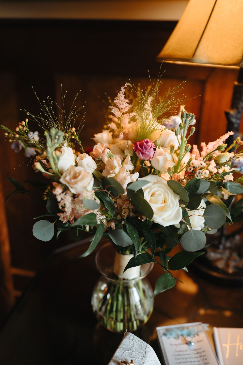 Beautiful wedding bouquet featuring white roses, pink flowers, and eucalyptus in a glass vase with warm lamp light.