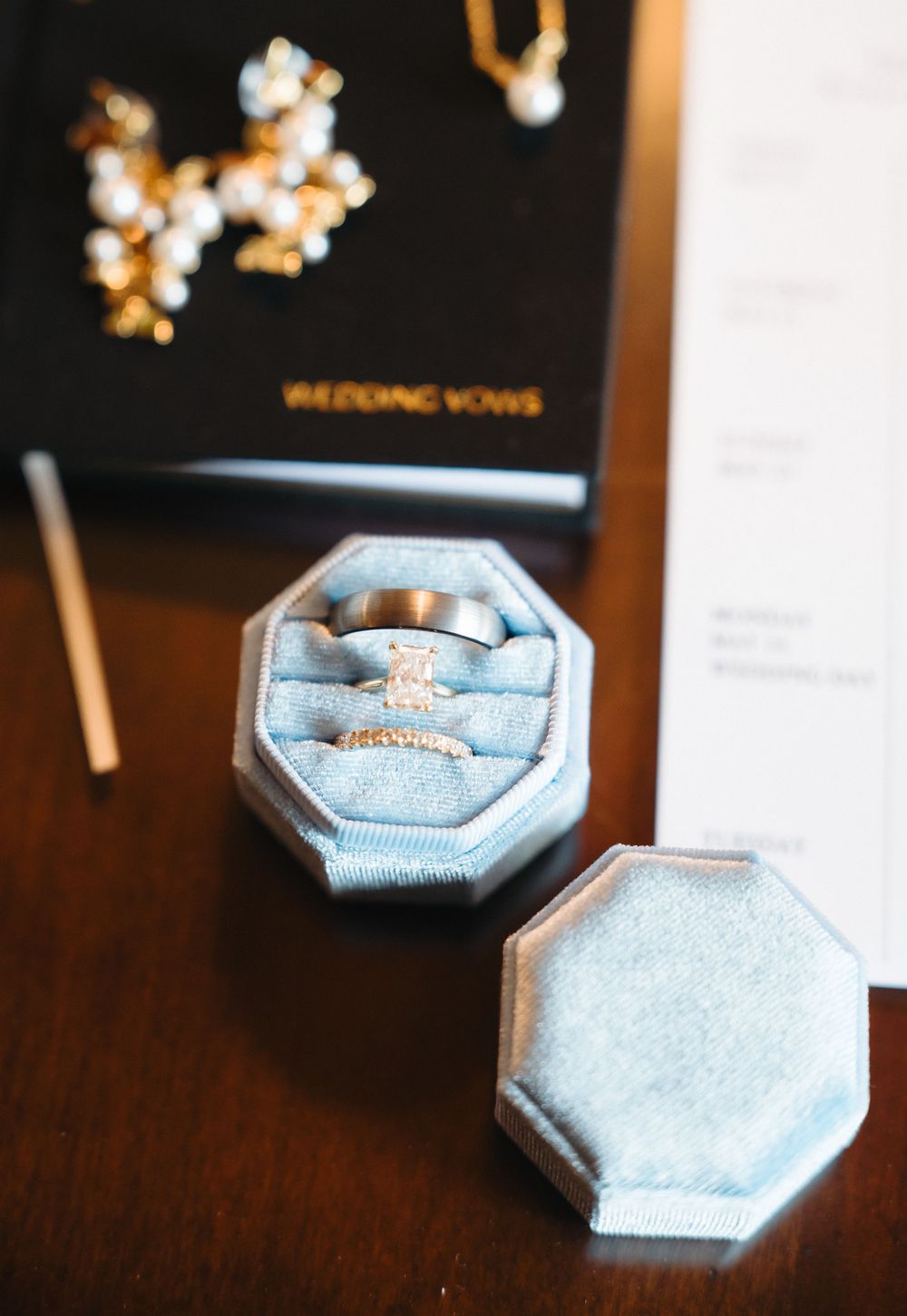 Blue velvet octagonal ring box with wedding bands and diamond engagement ring beside wedding vows booklet.
