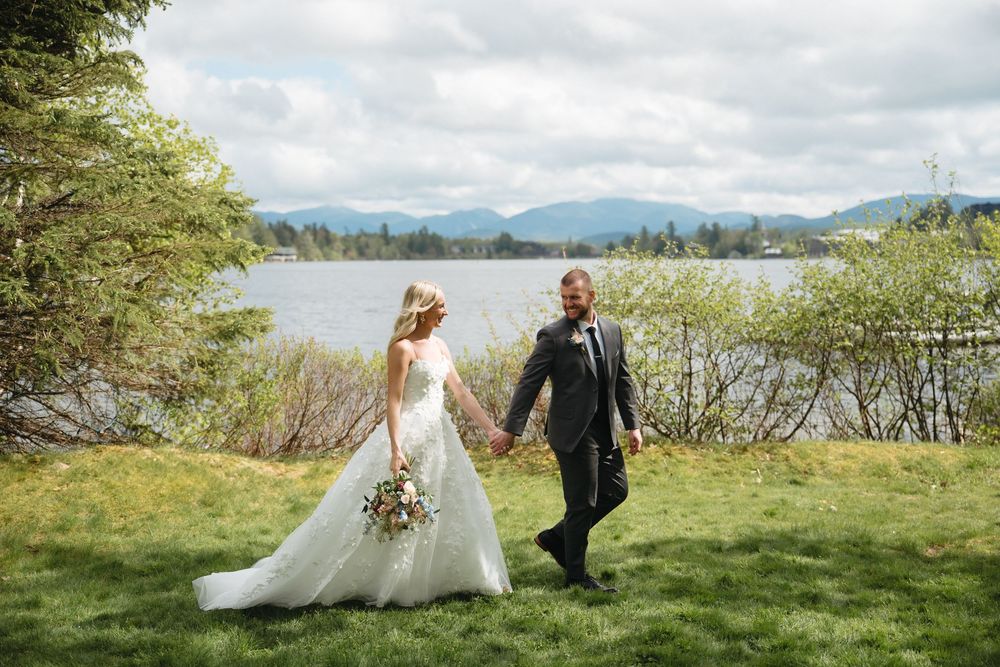 Bride and groom holding hands walking on green lawn with scenic lake and mountain backdrop on their wedding day.