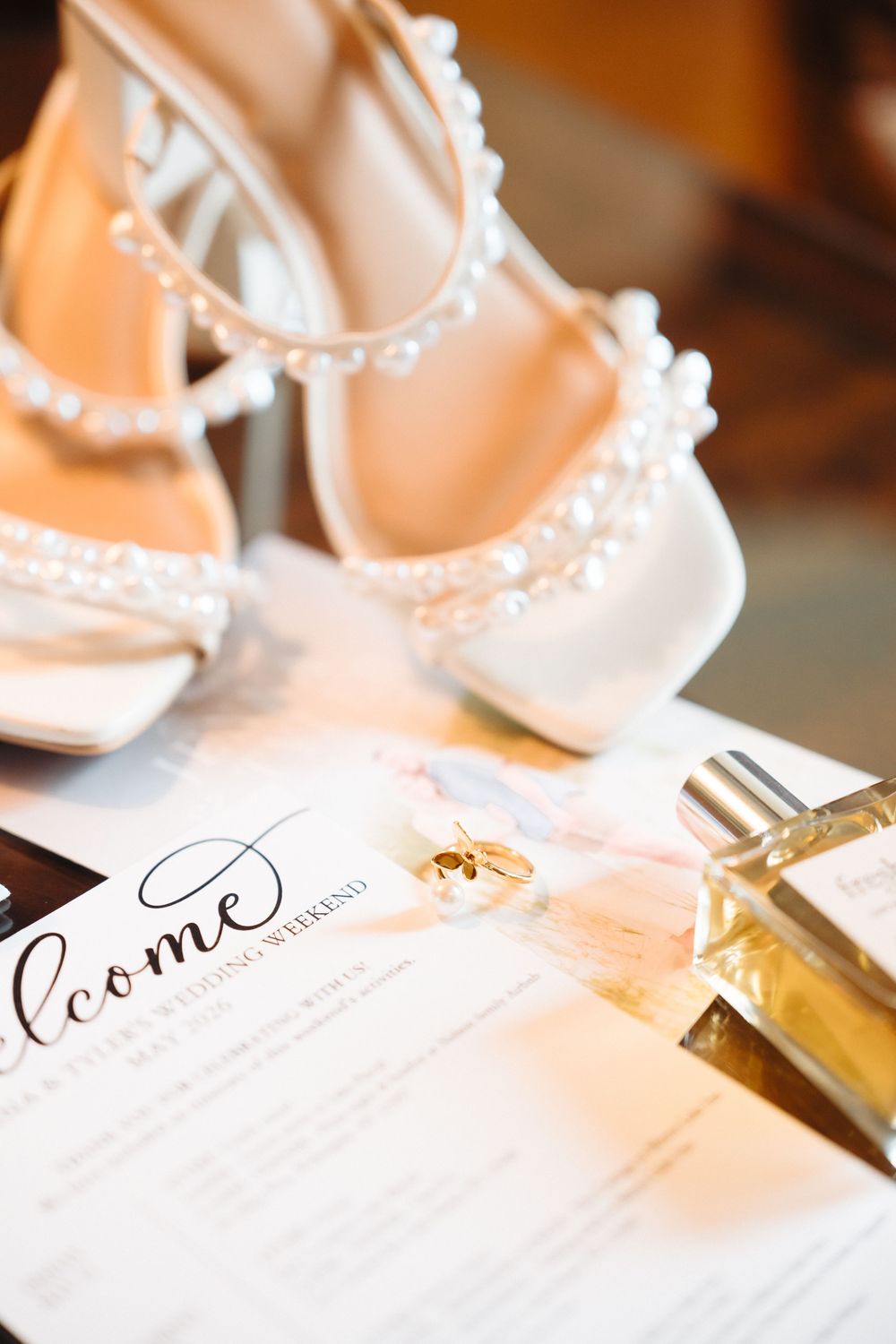 Close-up of pearl-trimmed white bridal shoes beside gold wedding rings, perfume bottle, and elegant welcome card.