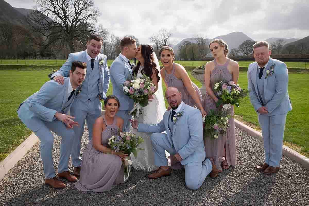 Wedding party posing outdoors with mountains, groomsmen in light blue suits and bridesmaids in taupe dresses holding floral bouquets.