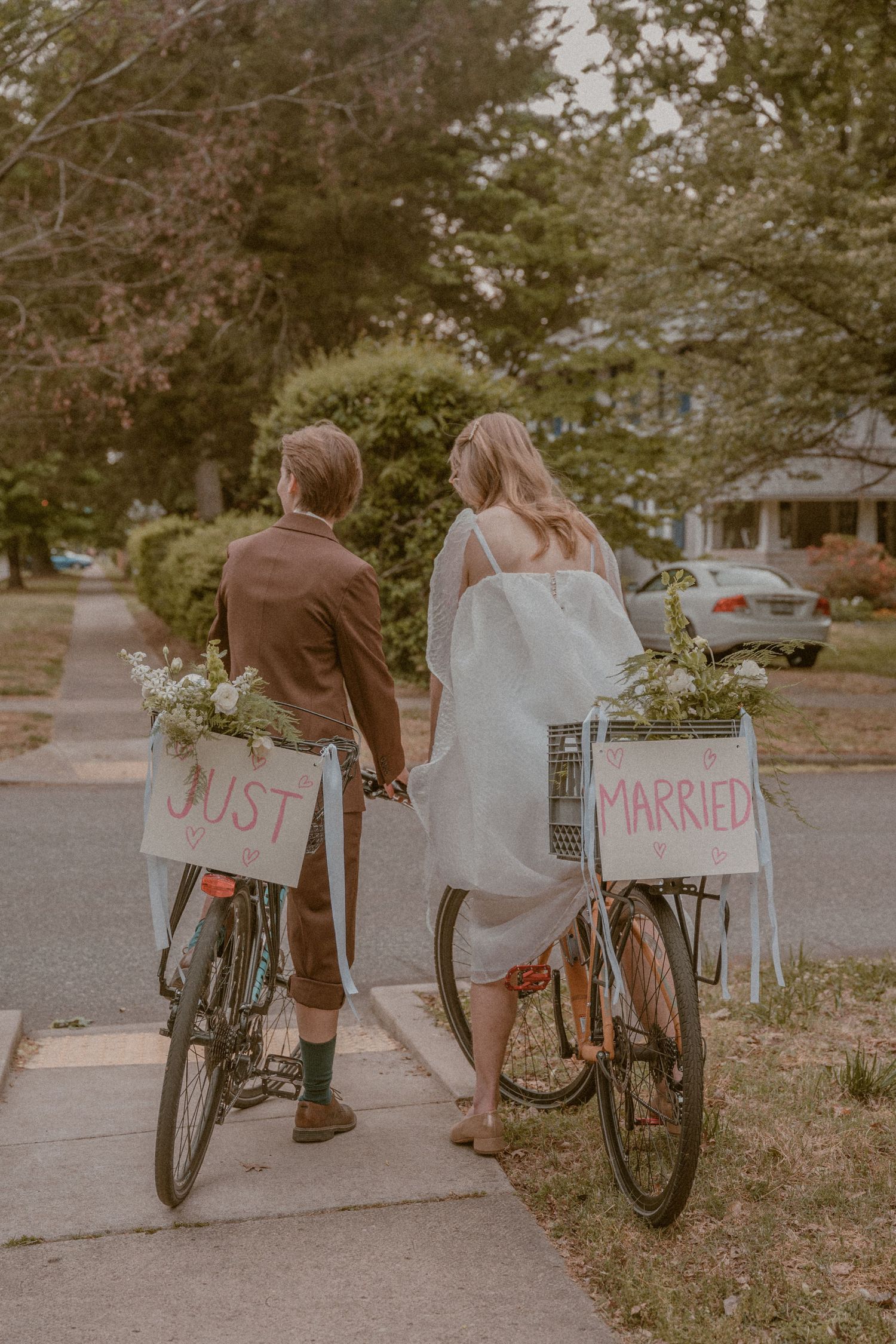 Newlywed couple walking bicycles with Just Married signs and flowers, holding hands on a suburban sidewalk.
