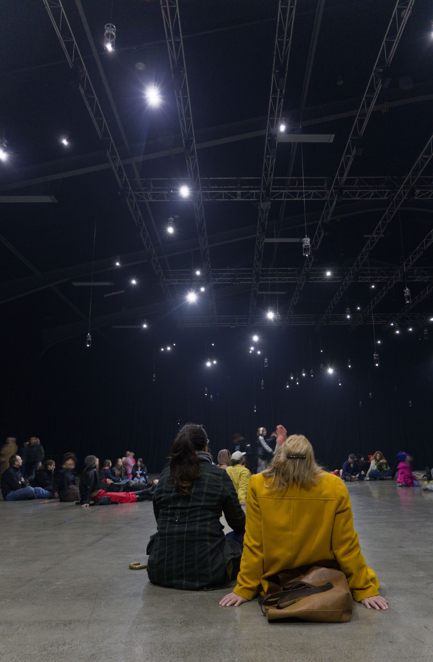 Audience members sit on the floor of a large dark venue watching a performance under dramatic stage lighting.