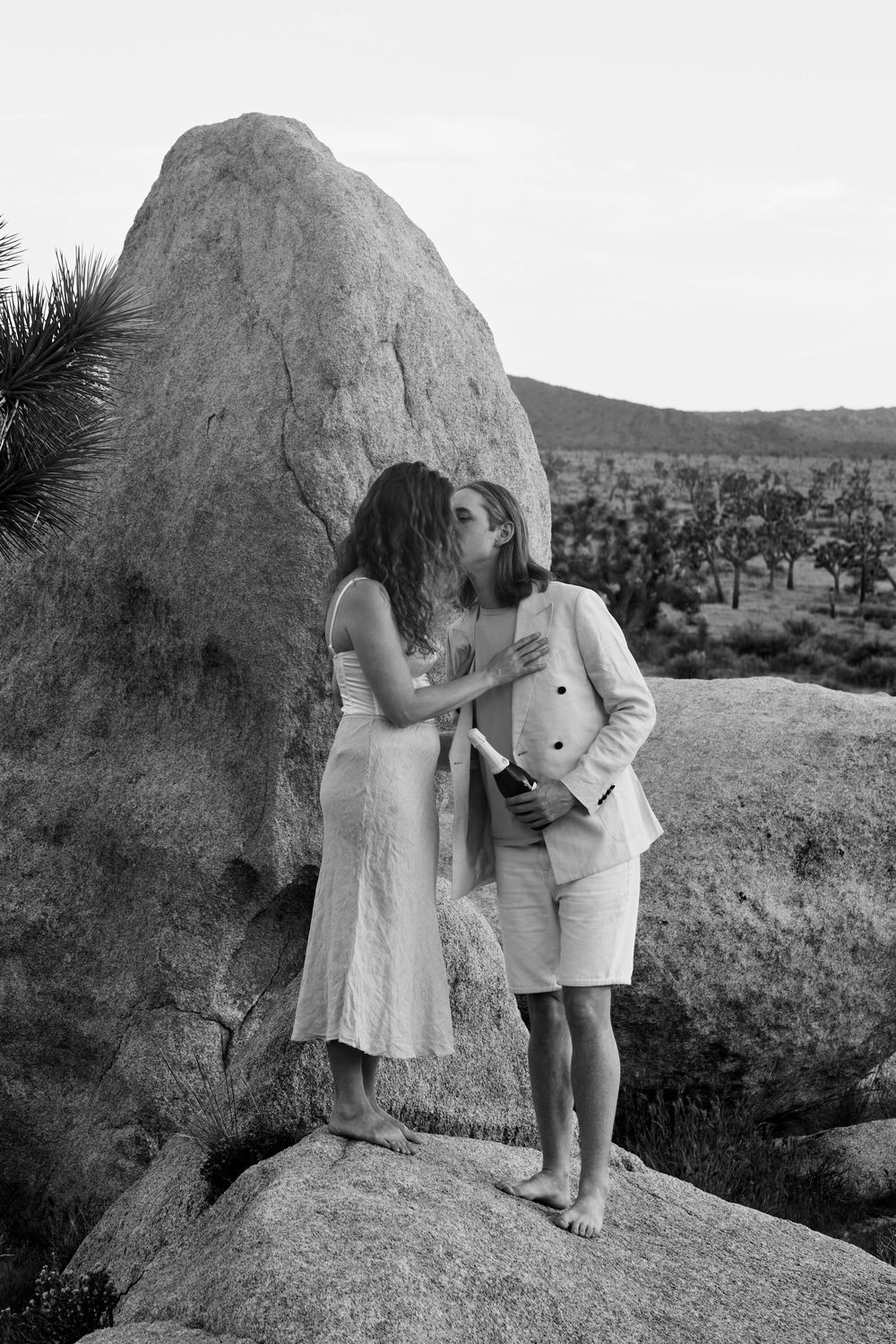 Black and white photo of a couple kissing on rocks in Joshua Tree desert, woman in white dress, man in light suit holding champagne.