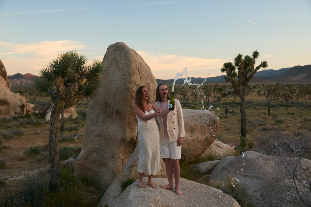 Two women in white dresses pose on desert rocks at sunset in Joshua Tree National Park.