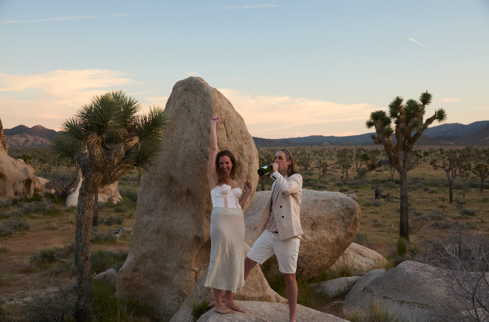 Two women in white outfits pose on desert rocks among Joshua trees at sunset in Joshua Tree National Park.