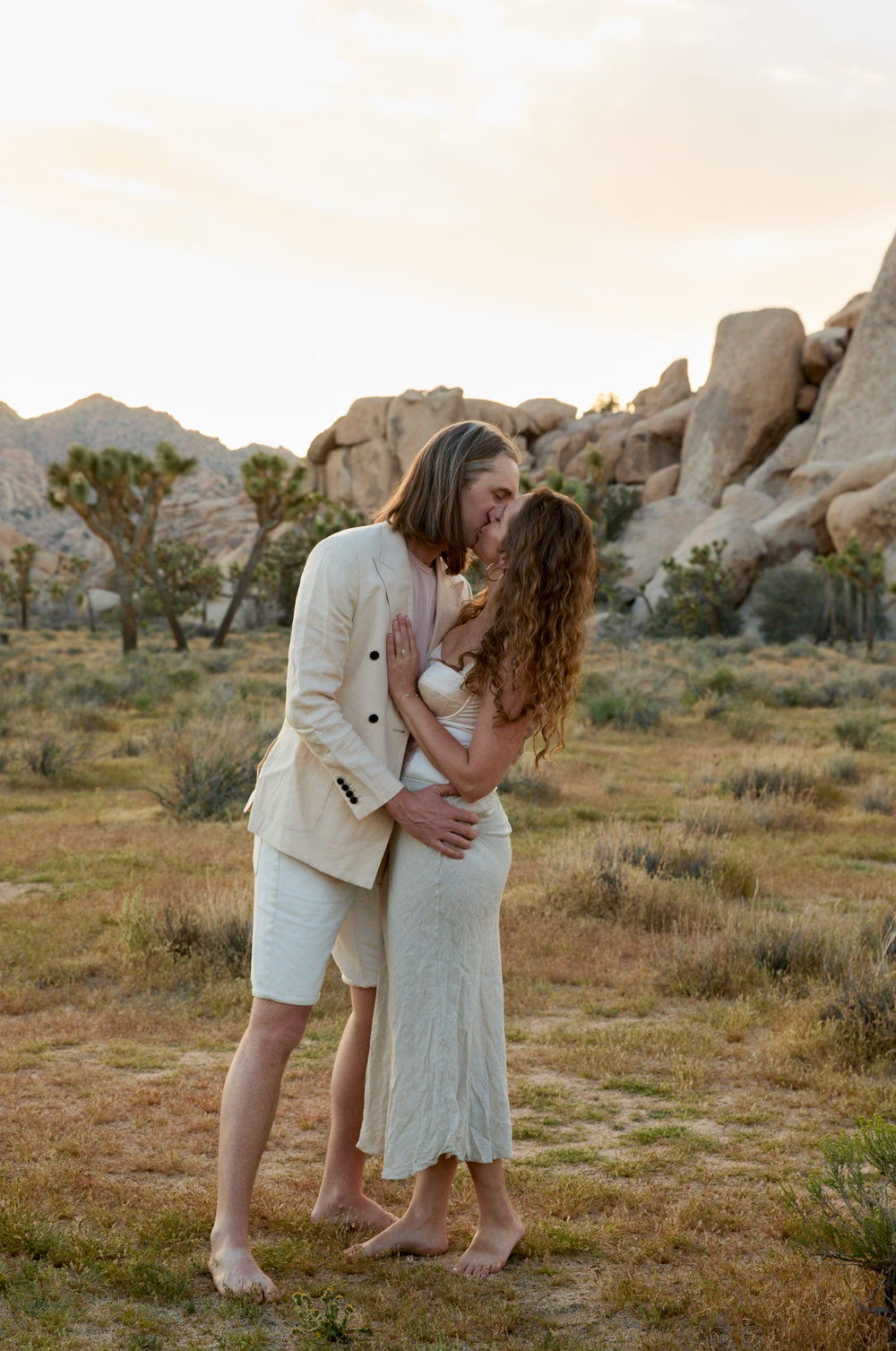 Couple sharing a romantic kiss at golden hour in Joshua Tree desert landscape with boulders and shrubs.