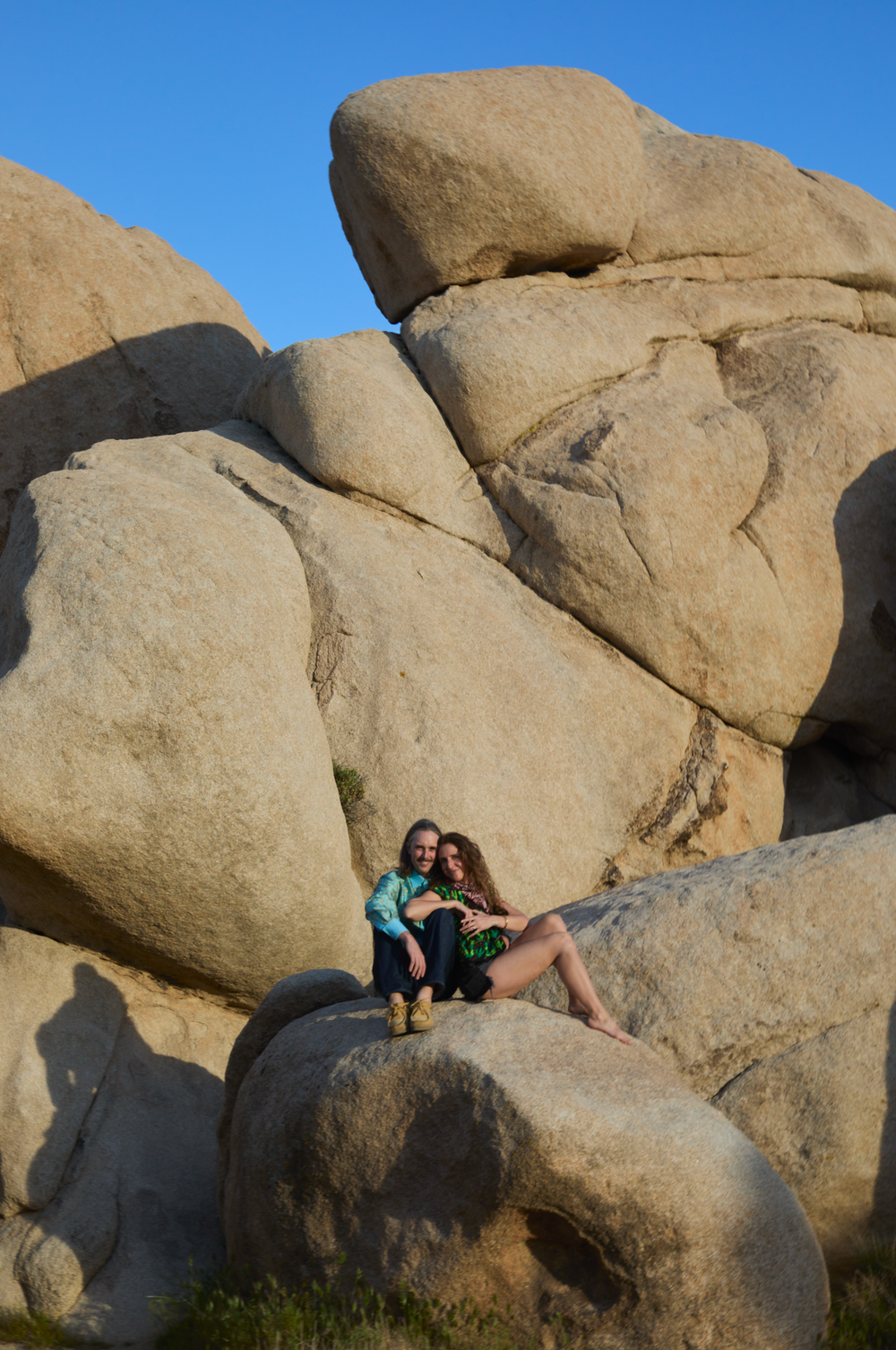 Couple sitting together on large granite boulders under a clear blue sky in a rocky desert landscape.