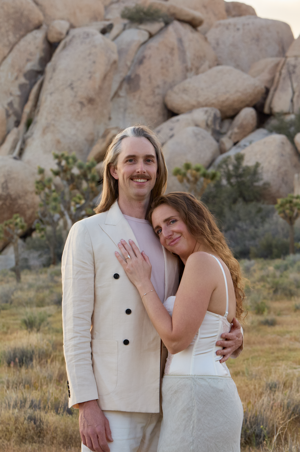 Couple in white wedding attire embracing in Joshua Tree desert landscape with large boulders and cacti.