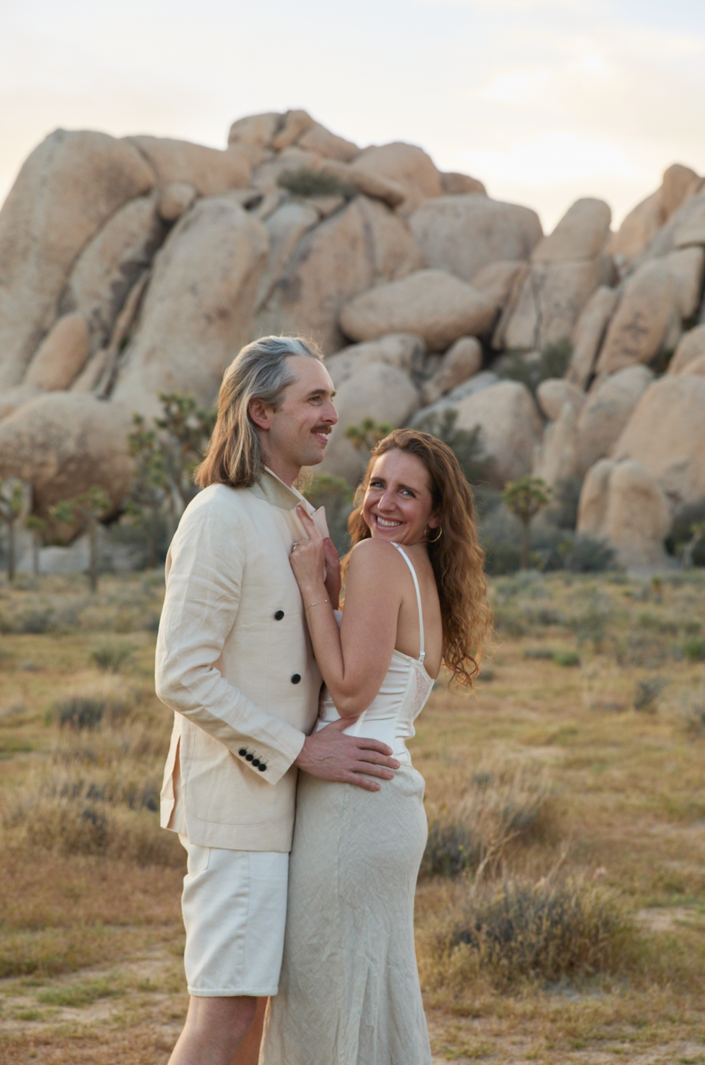 A couple in wedding attire embraces outdoors in Joshua Tree desert landscape with large boulders at sunset.