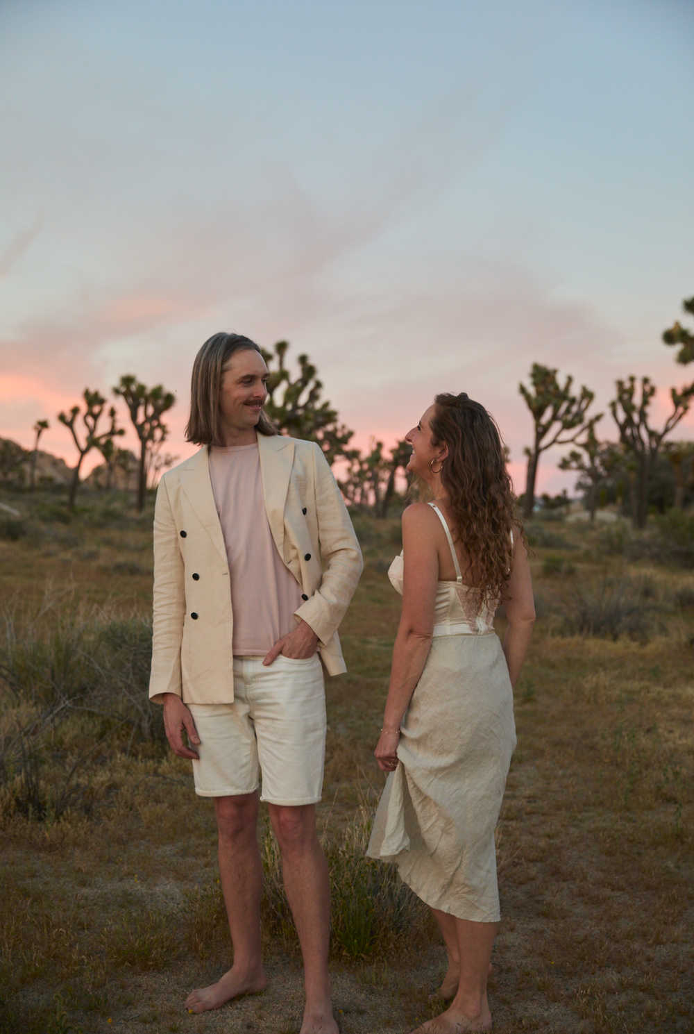 Couple in cream outfits standing in Joshua Tree desert at sunset, sharing a romantic glance.