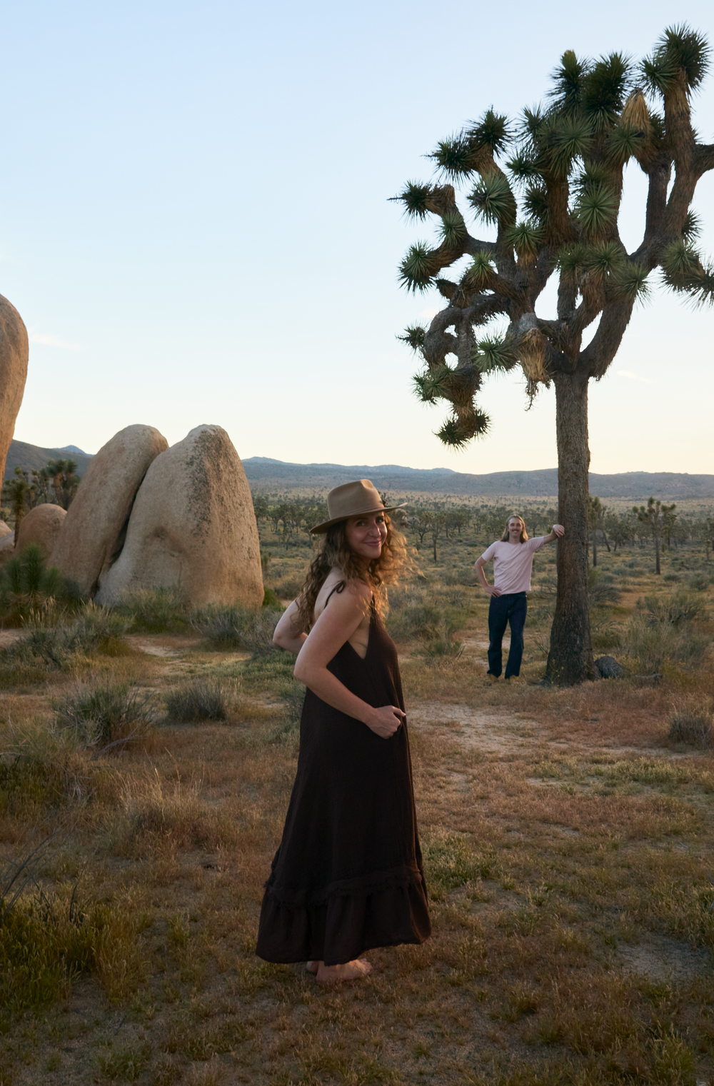 Woman in brown maxi dress and hat stands barefoot in Joshua Tree desert landscape at golden hour.
