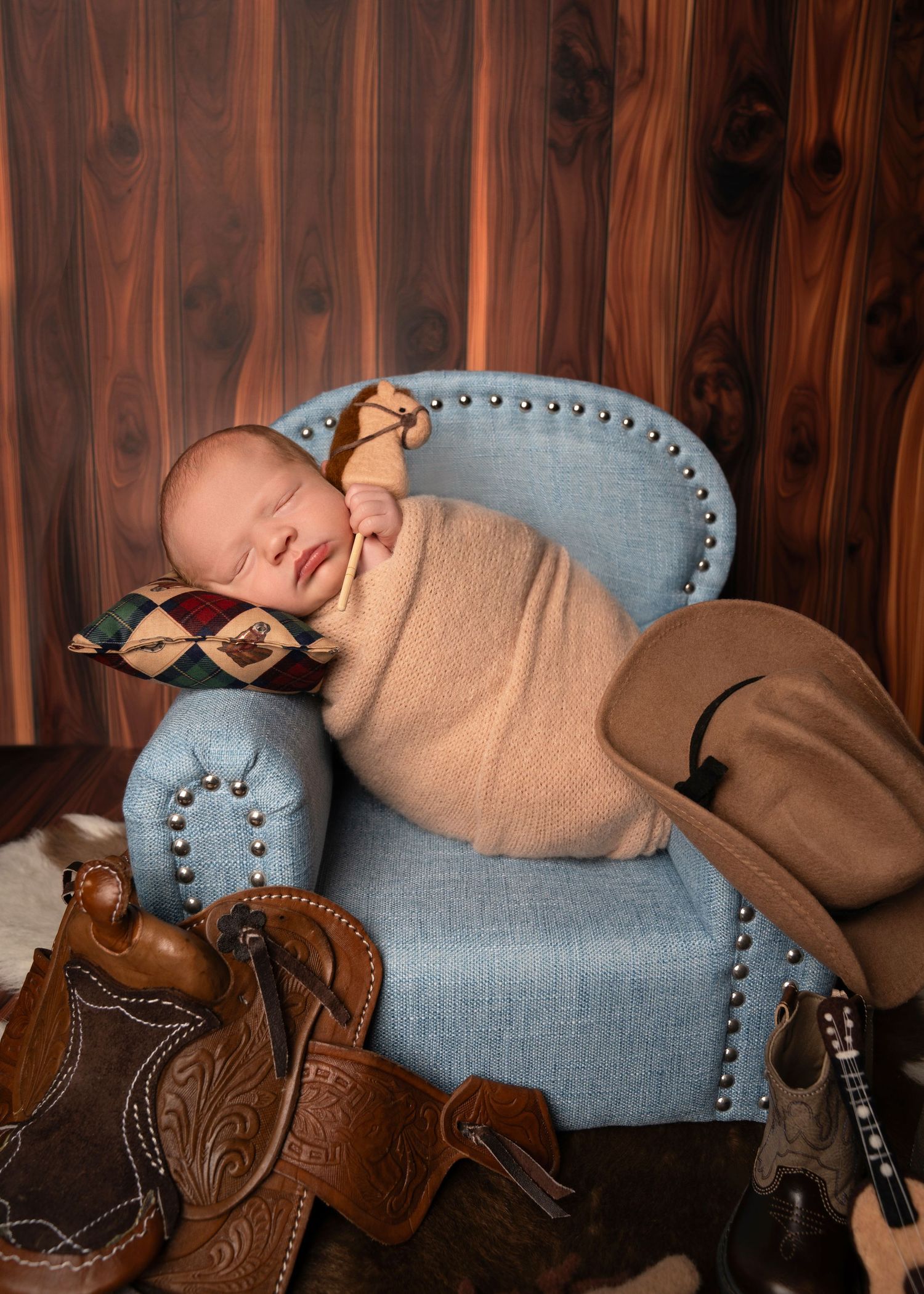 Sleeping newborn baby swaddled in tan wrap on blue tufted mini chair with cowboy saddle and boots in western-themed photo.